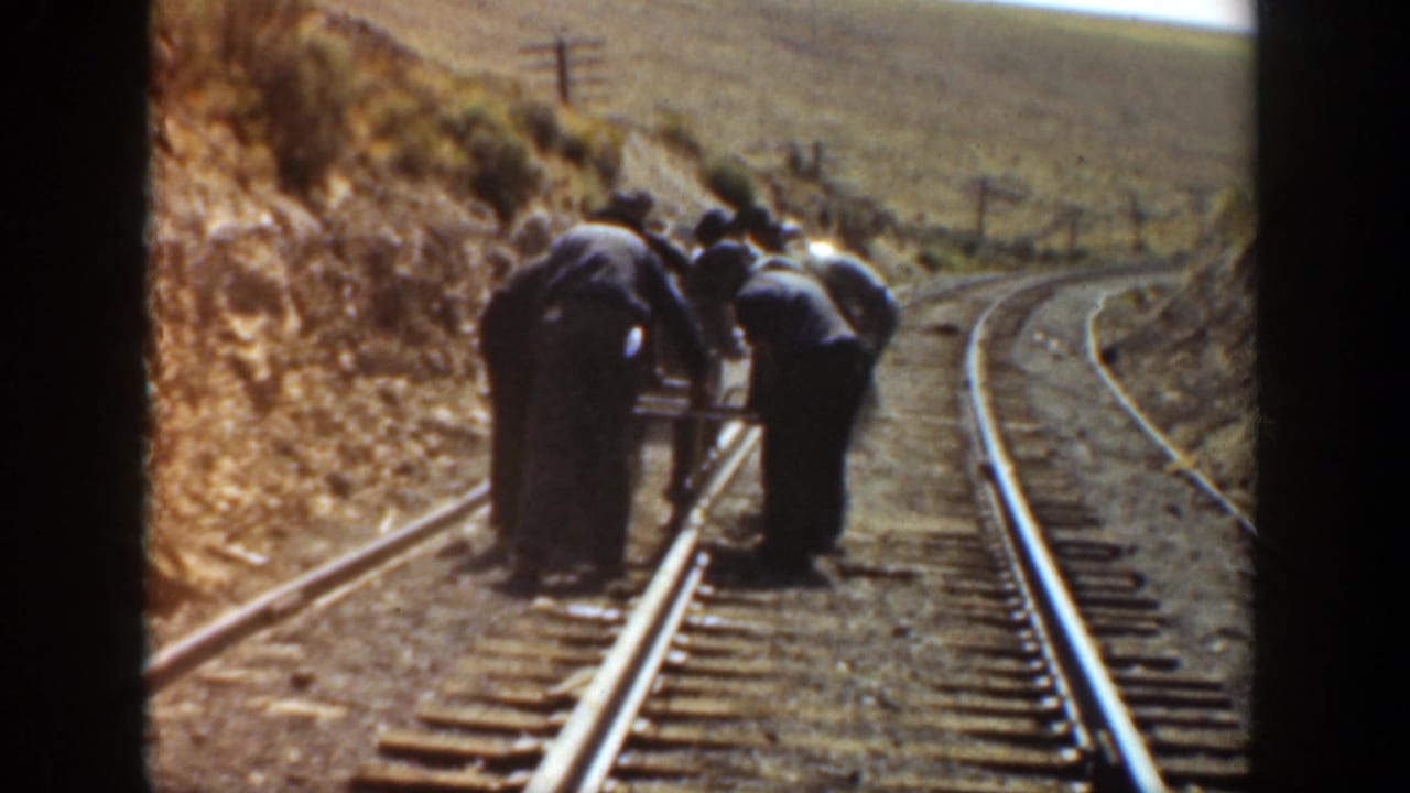 Workers in dark clothing bend over railroad tracks in a dry hilly landscape.