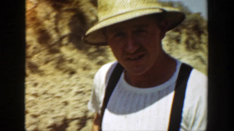 A man in a straw hat, white shirt, and dark suspenders appears close to the camera near the rail work site in California.