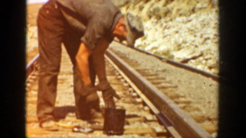 A worker bends beside the rail, with tools and a small container visible on the ties in the dry trackside setting.