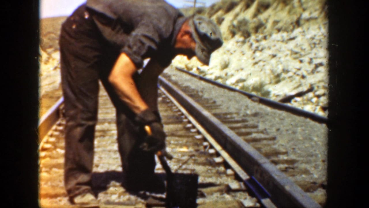 Man in a grey cap bends over railroad tracks with a tool beside a rocky slope.