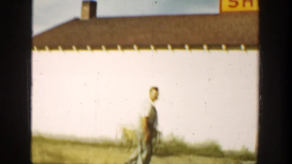 1936 California farmstead view with an adult man walking past a white building, brown roofline, and nearby wagon or equipment.