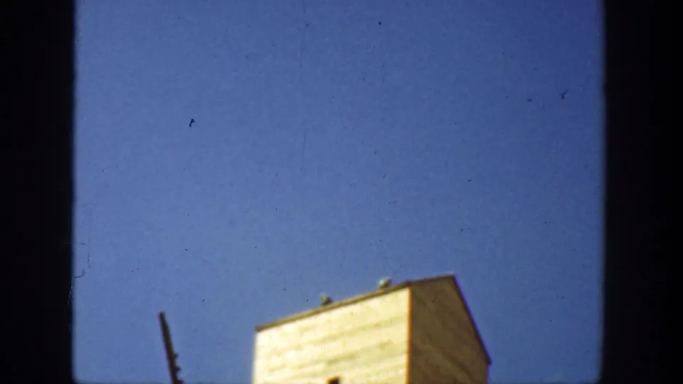 A light-colored California tower appears with small openings and a diagonal foreground pole or ladder in an archival 8mm frame.