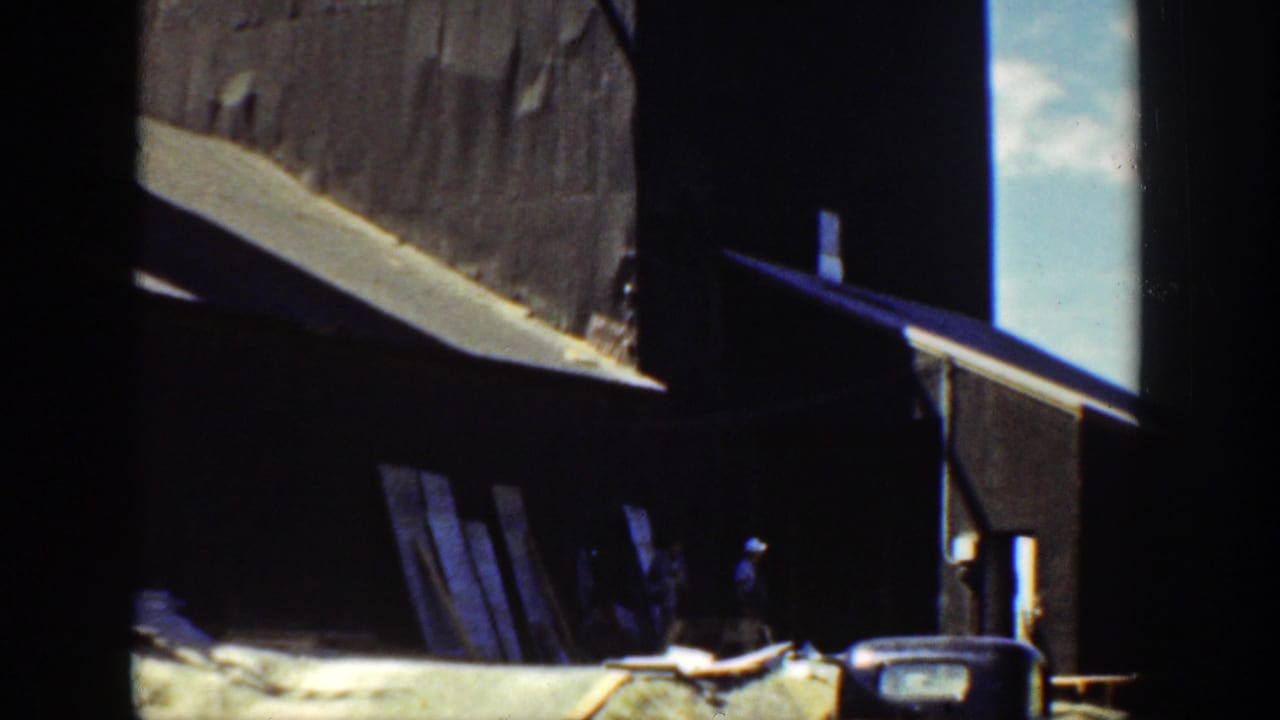 1936 California 8mm frame showing a shaded riverside work area with timber planks, containers, and bright daylight beyond.