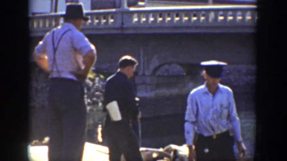 1936 California 8mm frame of workers beside a riverside wall, with period hats, suspenders, and sunlit work materials visible.
