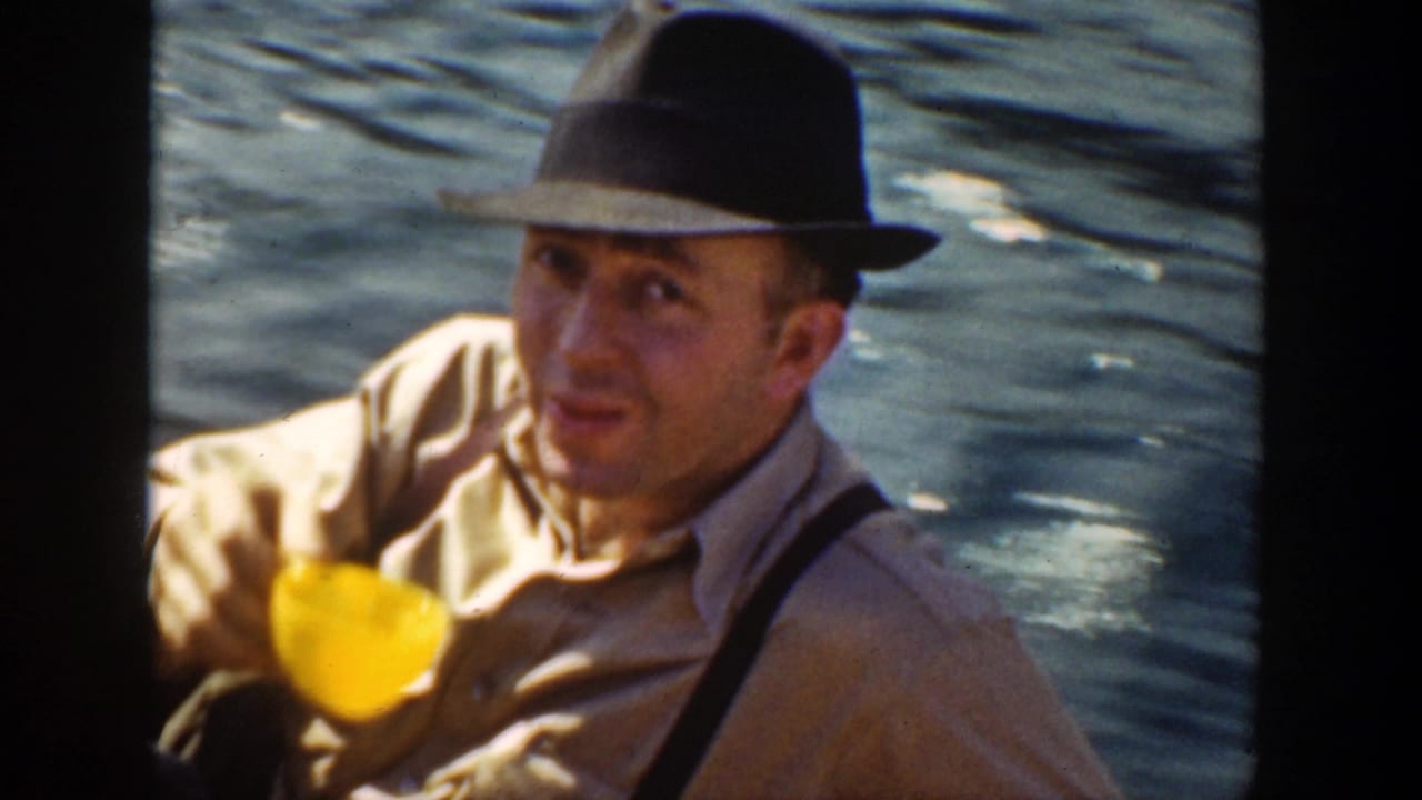 Man in a fedora holds a yellow cup and looks toward the camera by the water.