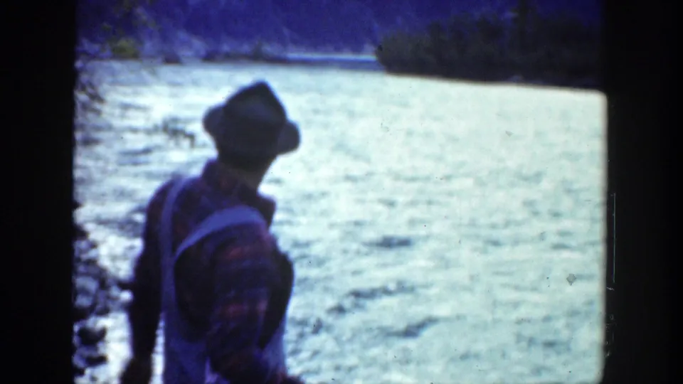 A man in a hat and suspenders stands on a rocky riverbank, facing the water during a 1936 California fishing outing.