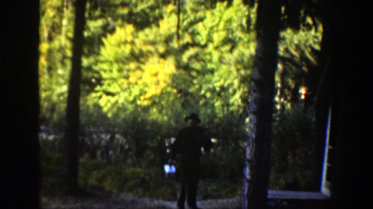 Man in dark clothing walks through trees carrying fishing gear.