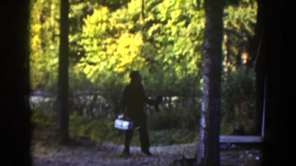 The man pauses or turns on a wooded California trail, holding a tackle box while framed by tree trunks and sunlit leaves.