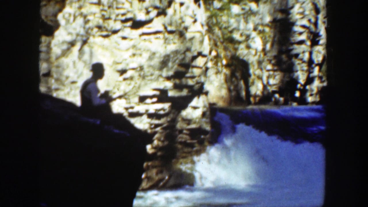 Man in a light shirt and hat sits on rock beside a rushing waterfall in a canyon.