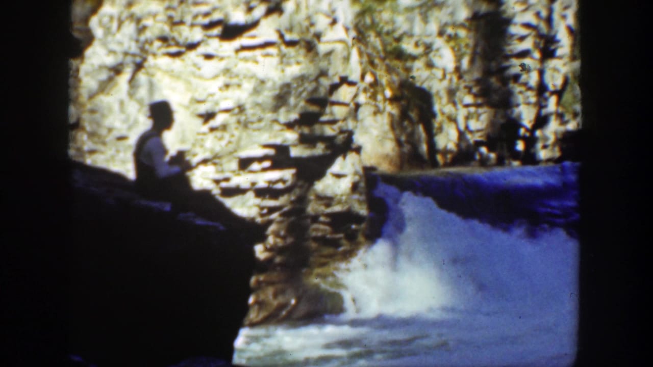 Seated man faces a waterfall as white spray rises below sunlit canyon rock.