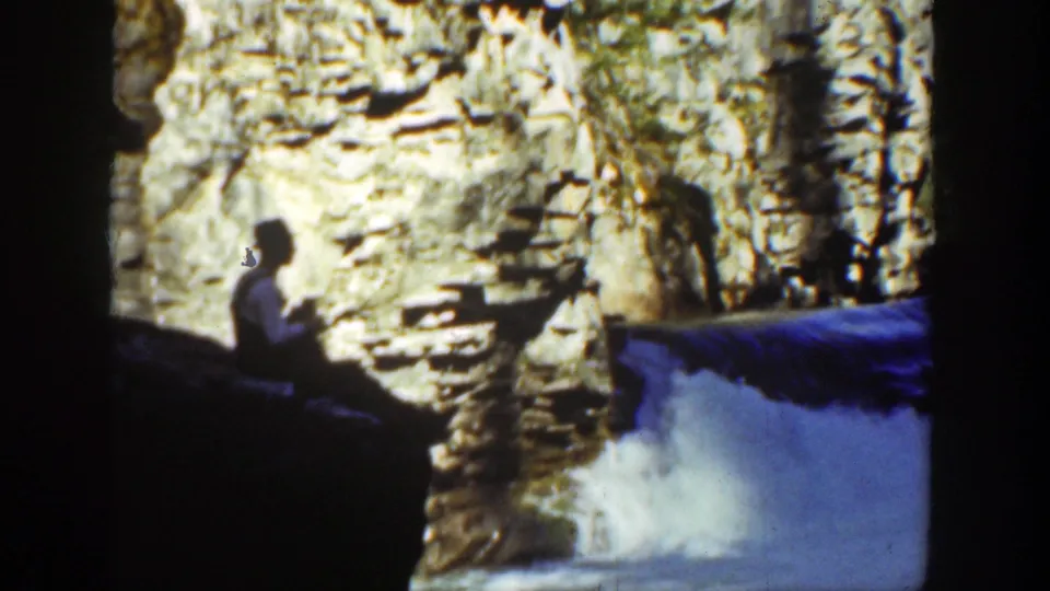 1936 California 8mm still of a man seated in shadow beside a rugged canyon wall, looking toward a bright waterfall and blue water.
