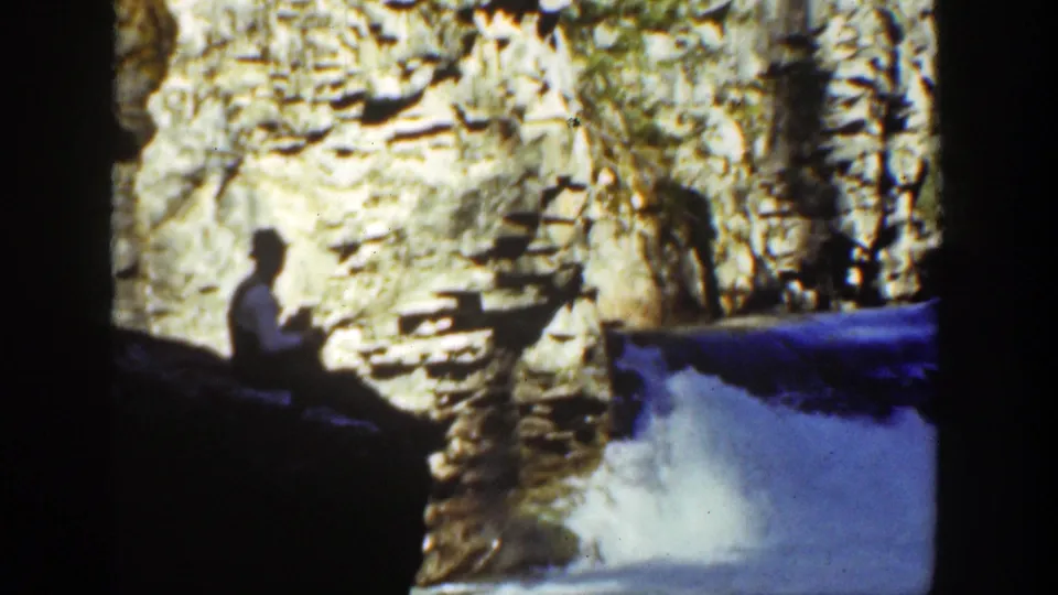 1936 California home-movie frame with a seated man near a cliff face, waterfall spray below, and strong contrast between shade and sunlit rock.