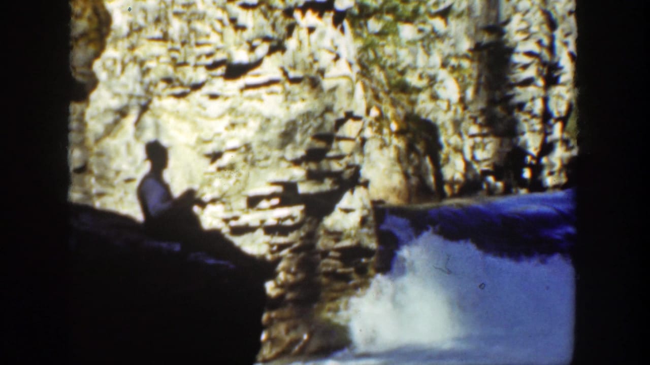 Man in a hat sits beside a rocky waterfall with foaming water below.