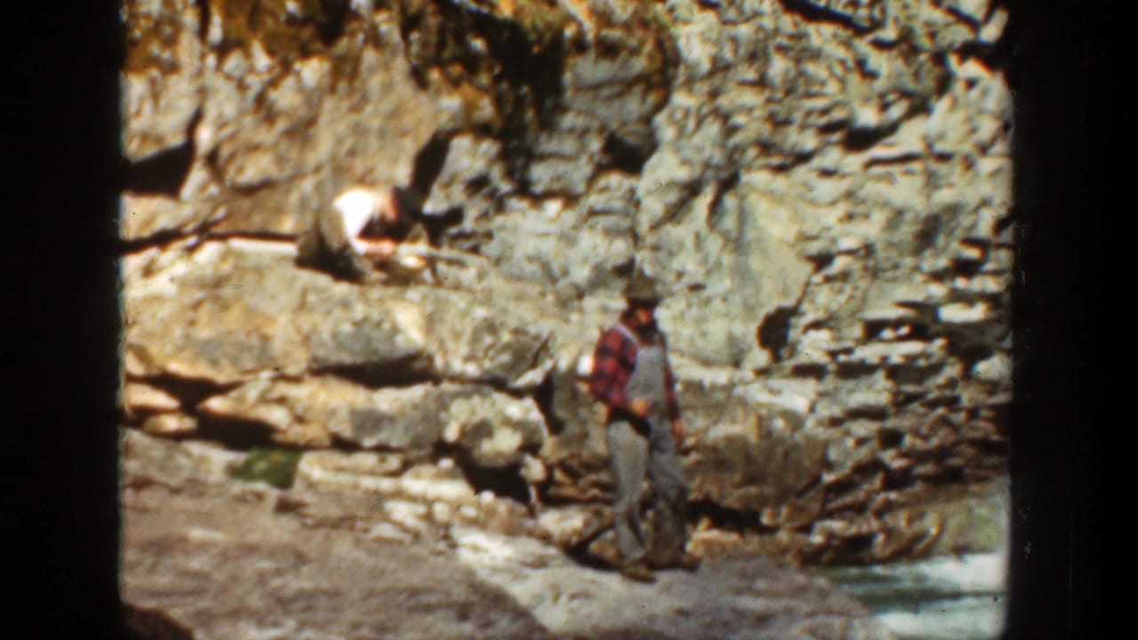 Two men near layered canyon rocks, one standing by the riverbank and one seated above.