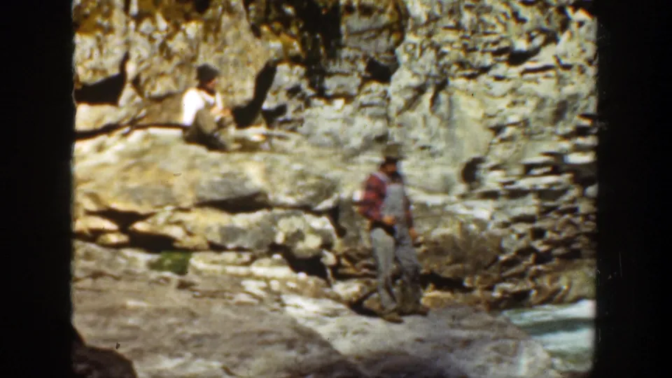 1936 California canyon still with a standing man in work clothes below a seated man on layered rock beside water.