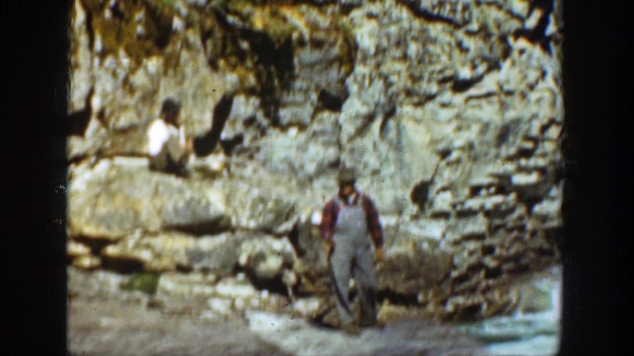Man in red shirt and overalls stands in front of a rocky canyon wall near water.