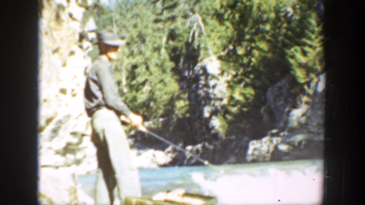 Man in a brimmed hat stands beside a river holding a fishing rod.