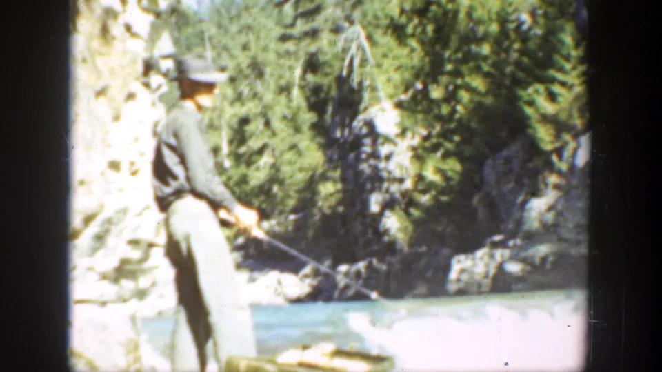 1936 California frame of an adult man in light shirt and trousers fishing from a rocky riverbank among trees.