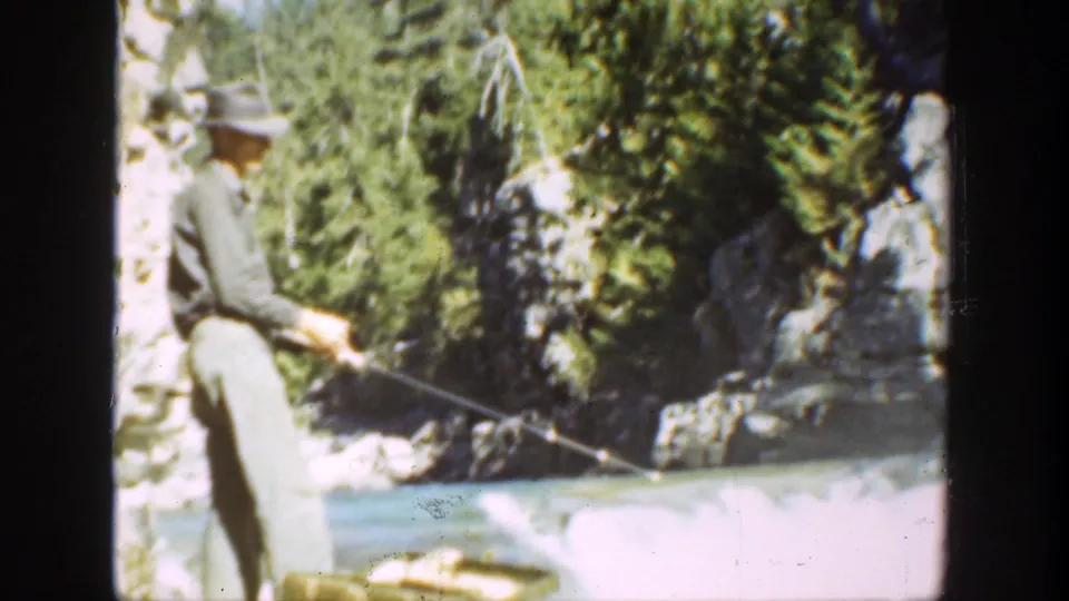 Adult man in 1936 California home-movie footage casts or holds a fishing line at a fast river bordered by rocks and evergreens.