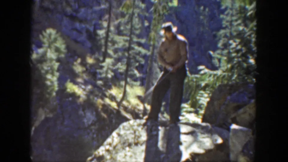 The golfer stands on a boulder in a California mountain forest, with tall evergreens and rocky slopes behind him.