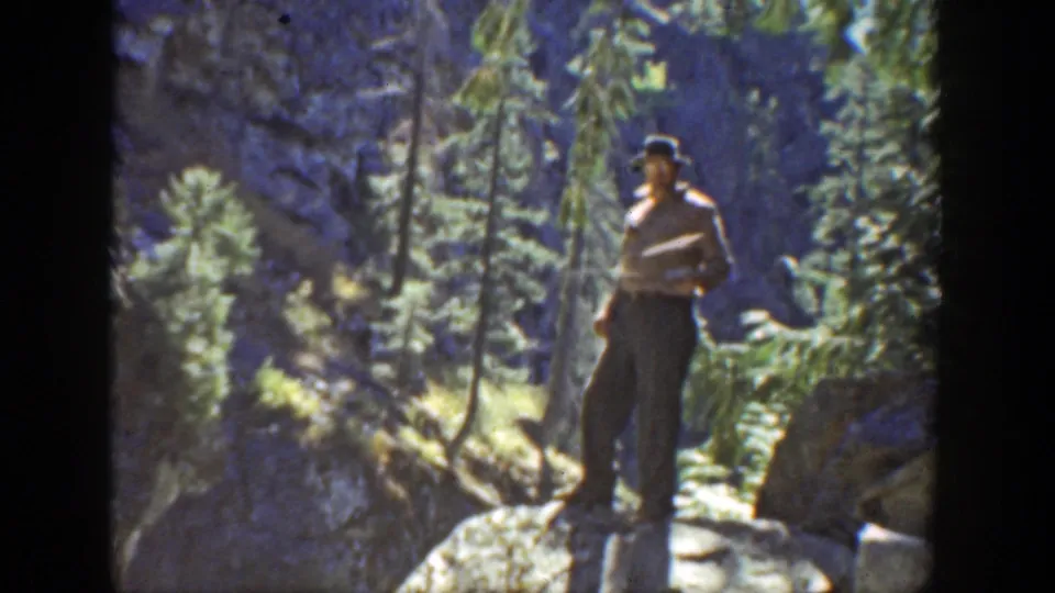 A man in 1930s California footage faces the camera with a golf club while standing on rocks in a rugged evergreen setting.