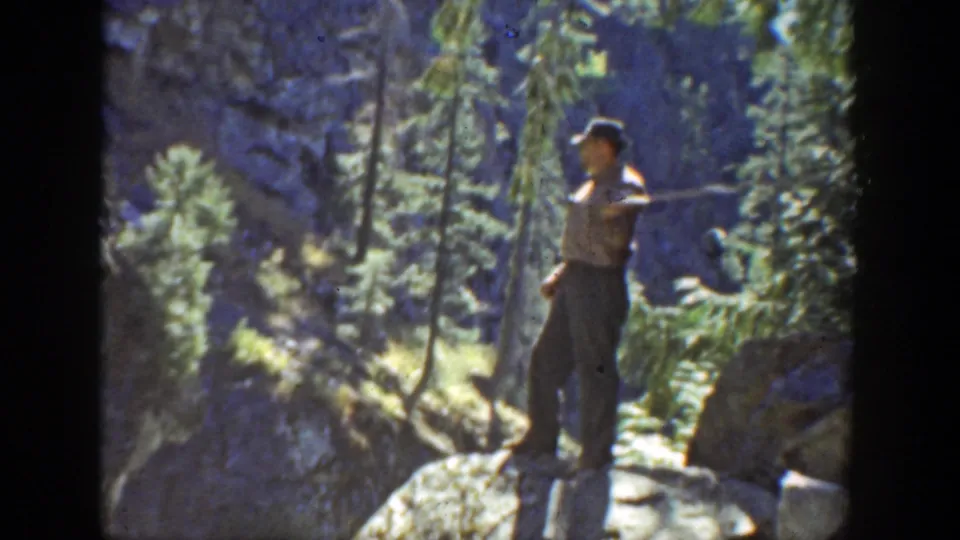 The man appears to extend or swing a golf club from a rocky perch in the 1936 California mountain forest footage.