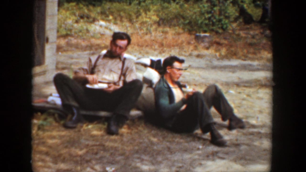 Two adult men in 1936 California home-movie footage sit on the dirt outside a building, one wearing suspenders and the other a dark sweater.