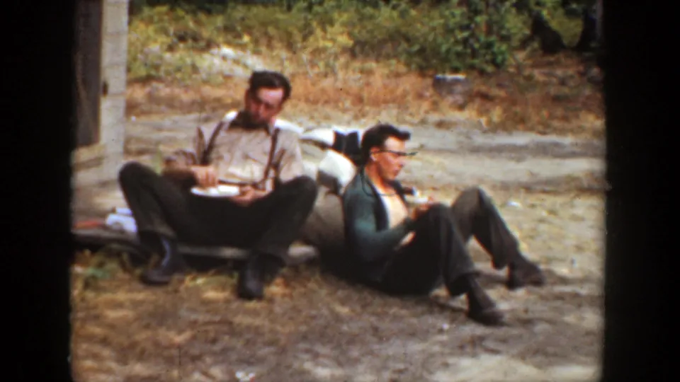 Two adult men in 1936 California home-movie footage sit on the dirt outside a building, one wearing suspenders and the other a dark sweater.