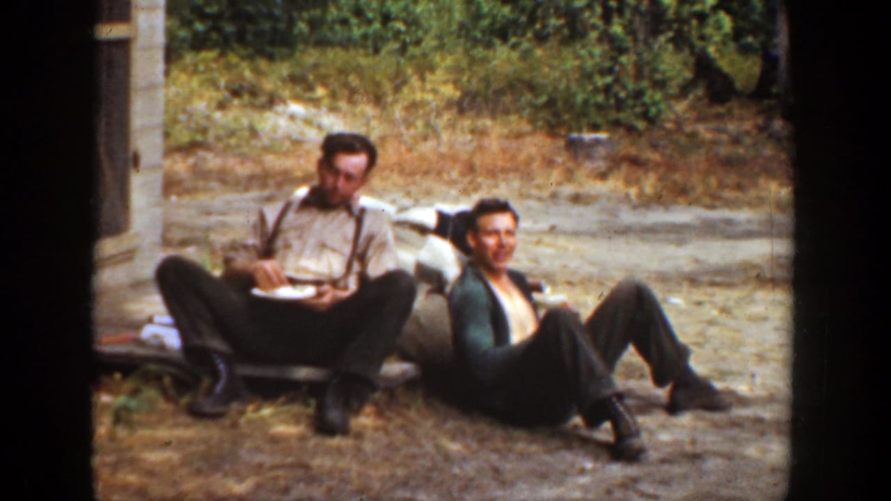 Two men sit on the ground with plates as one turns his face toward the camera.