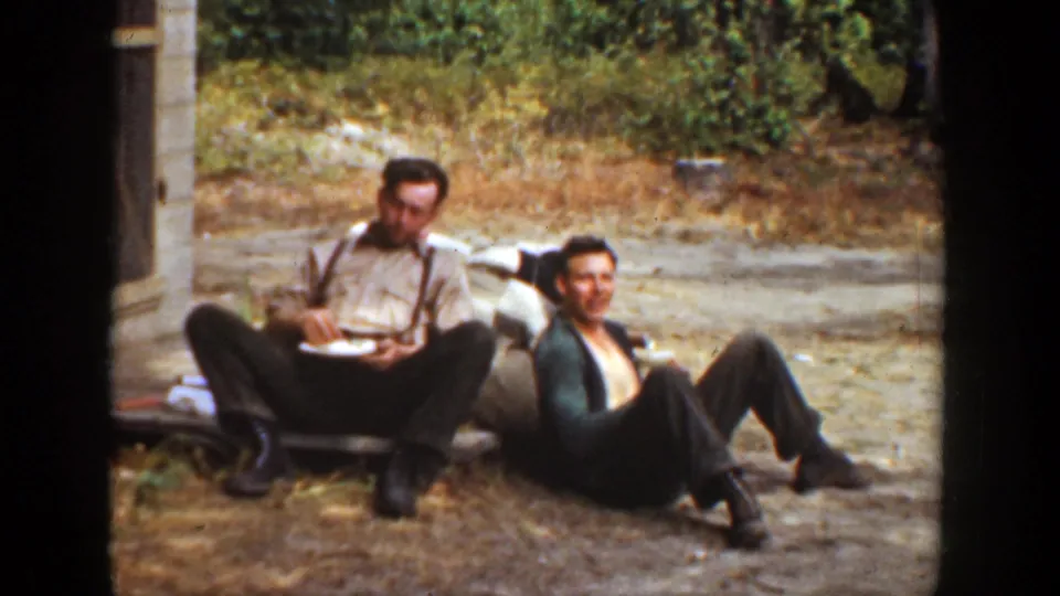 In this 1936 California frame, two men remain seated outdoors on dirt ground with plates, with foliage behind them and a building edge nearby.