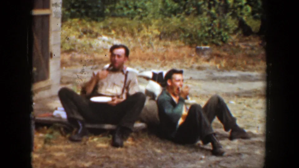 Two adult men in warm-toned 8mm footage sit low on the ground outside, eating from plates beside a building in California.