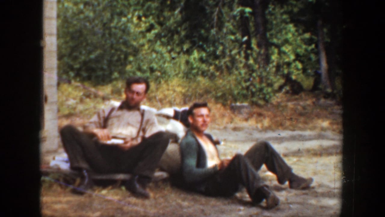Two adult men sit on the ground outdoors, one holding a plate while the other leans beside him.