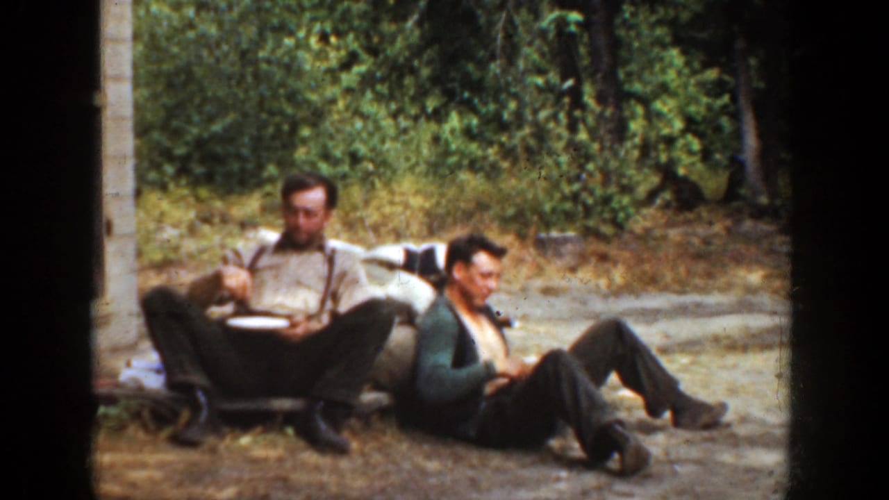 Two men remain seated outdoors as one looks down at a plate and the other turns slightly away.