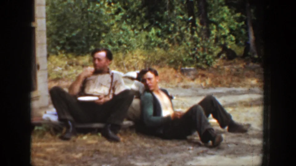 Two adult men sit close together outdoors in California, 1936, with one appearing to eat while the other looks toward the camera.