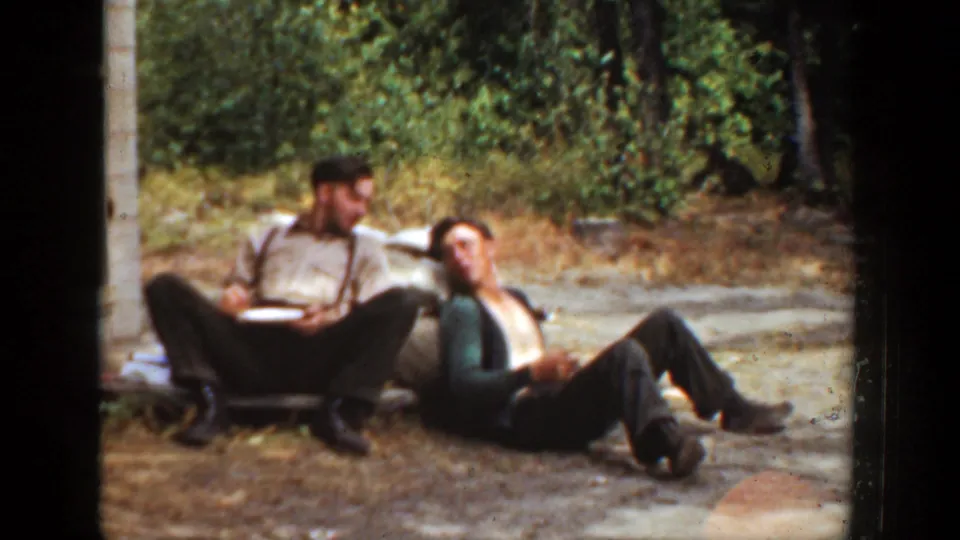 Two men in work clothes sit near trees in 1936 California, appearing relaxed as they turn toward each other during a meal break.