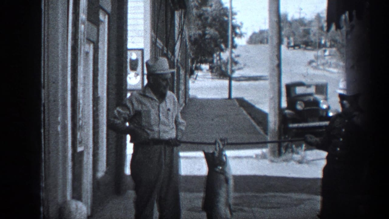 1936 California home-movie frame showing a man displaying a large fish on a sidewalk beside a storefront, with a uniformed officer partly visible.