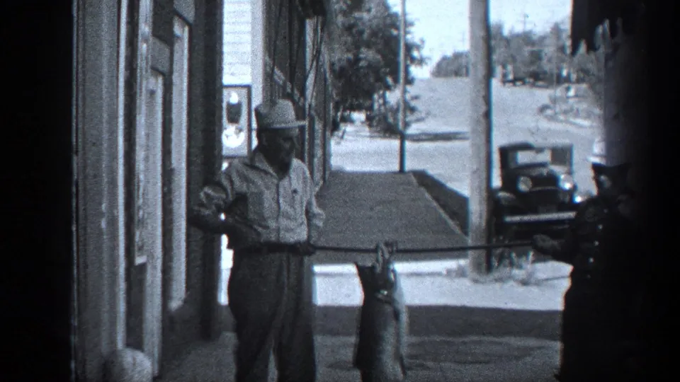 1936 California home-movie frame showing a man displaying a large fish on a sidewalk beside a storefront, with a uniformed officer partly visible.