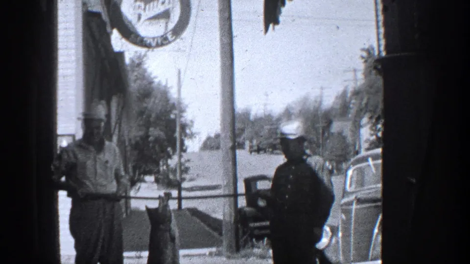 1936 California home-movie frame showing a sidewalk fish display between a man and a uniformed officer, with vintage vehicles nearby.