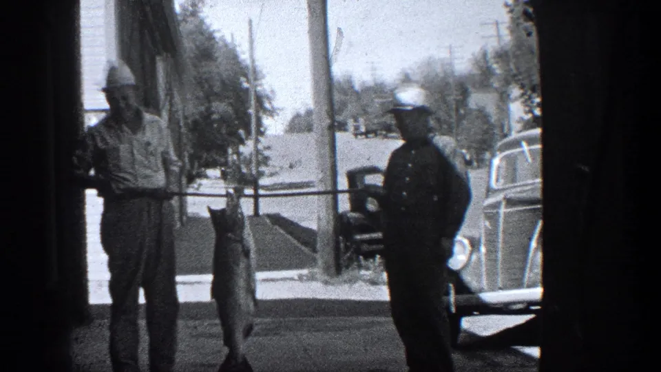 1936 California frame with a man displaying a large fish on a sidewalk beside a uniformed officer and vintage cars.