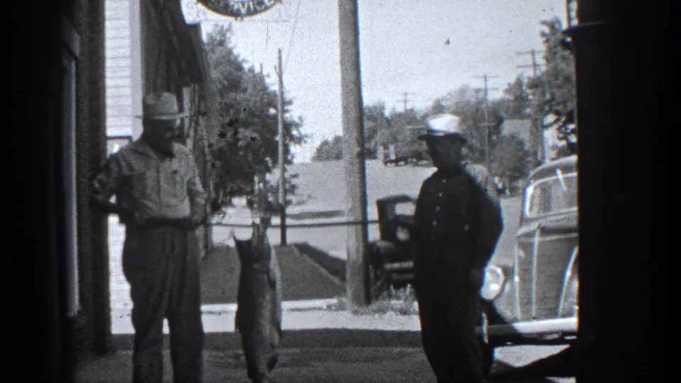 In 1936 California home-movie footage, two adult men in hats pose on a street with a large fish suspended from a pole.