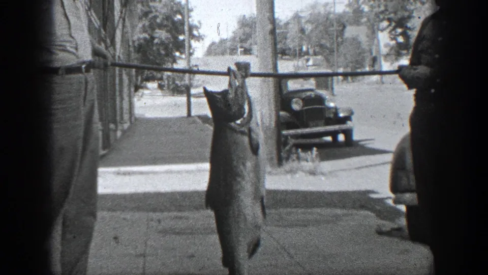 A large fish suspended from a pole dominates the frame, with a man partly visible and 1930s California street details behind it.