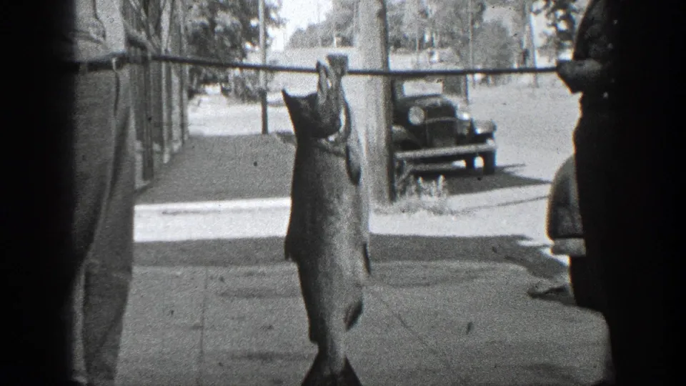 The home-movie frame centers on a large fish hanging from a pole, with a shaded sidewalk, parked car, and street in California.