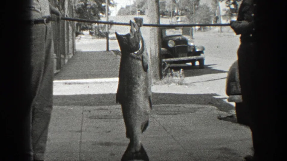 In 1936 California home-movie footage, two men display a large fish suspended from a pole beside parked cars and a sidewalk.