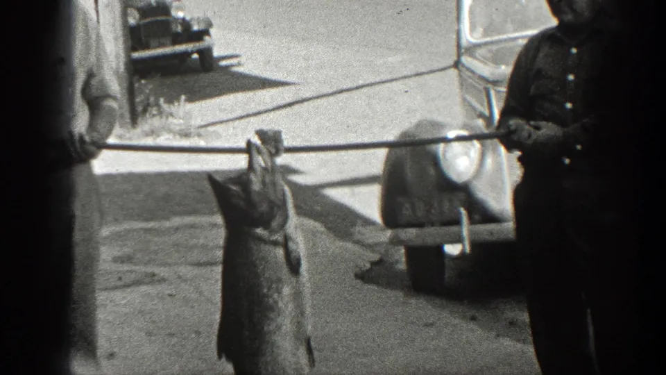A large fish hangs from a pole held by two men, with 1930s-era cars and a quiet street visible behind them.