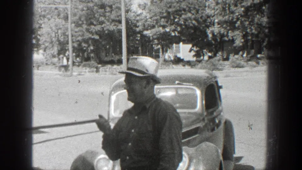 A man wearing a light hat stands beside a period car as the fish display pole passes across the foreground.