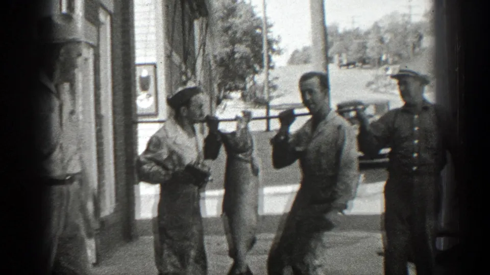 Men in period clothing stand beside a storefront as a large fish is held up between them on a pole.
