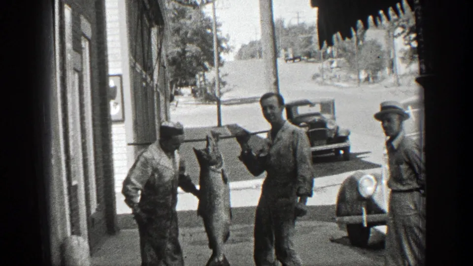 A sidewalk scene shows one man gesturing toward a large hanging fish as another man stands near a parked vintage car.