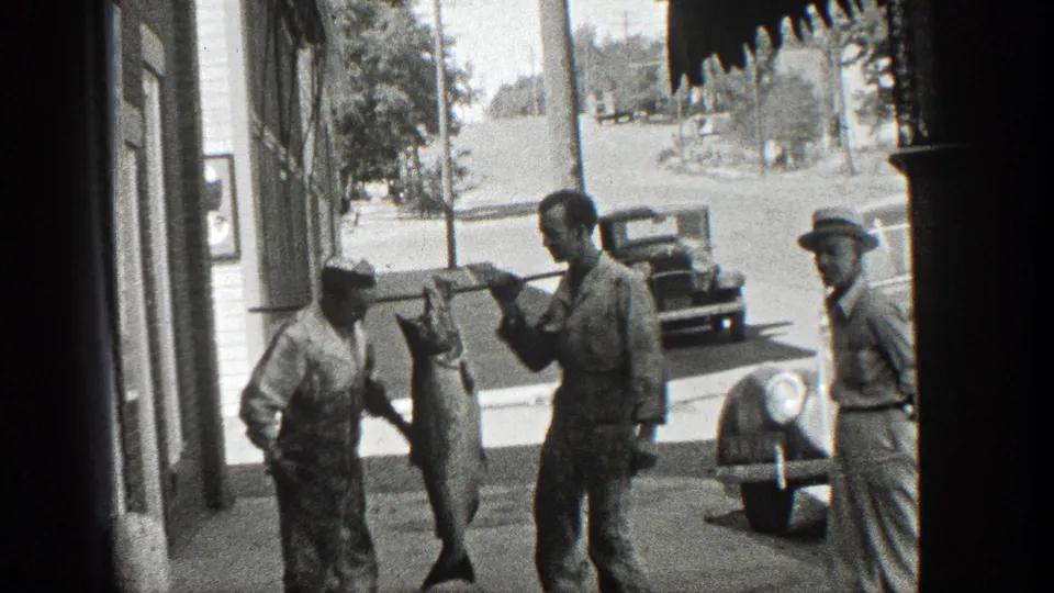 The large fish remains suspended from a pole as men pose on a California sidewalk with an early automobile behind them.