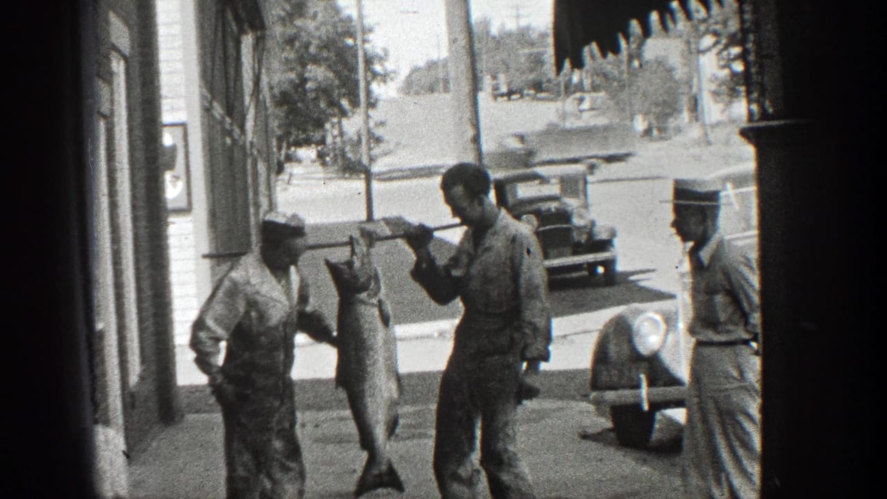 Men on a sidewalk display a large fish on a pole near parked vintage cars.