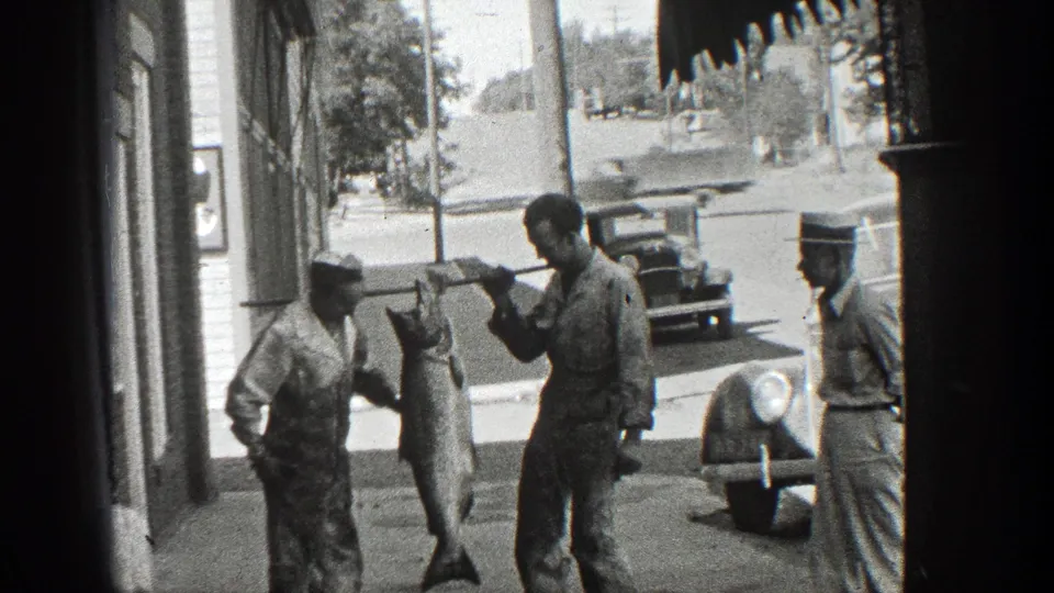 1936 California home-movie frame of men in work clothes and hats standing by storefronts as a large fish hangs from a pole.